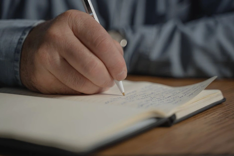 Osoba pisząca w notatniku przy biurku z laptopem.